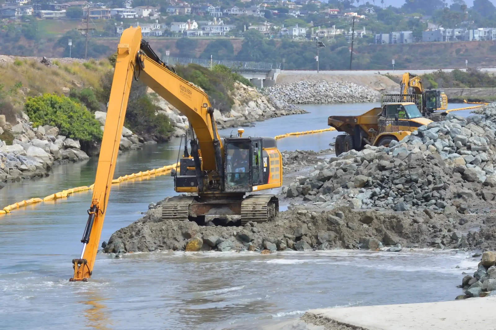 Yellow long-reach excavator dredging a waterway near rock piles and a coastal residential area.